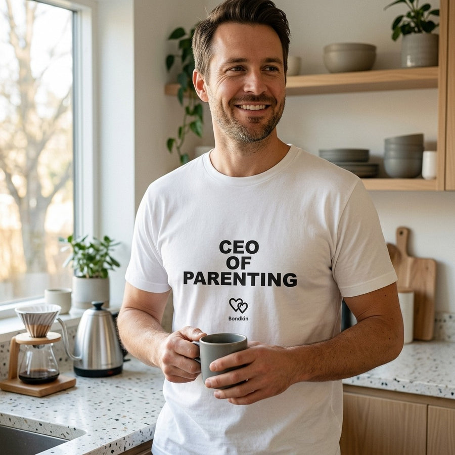 Smiling man in a white Bondkin CEO of Parenting tee holding a coffee mug in a bright kitchen